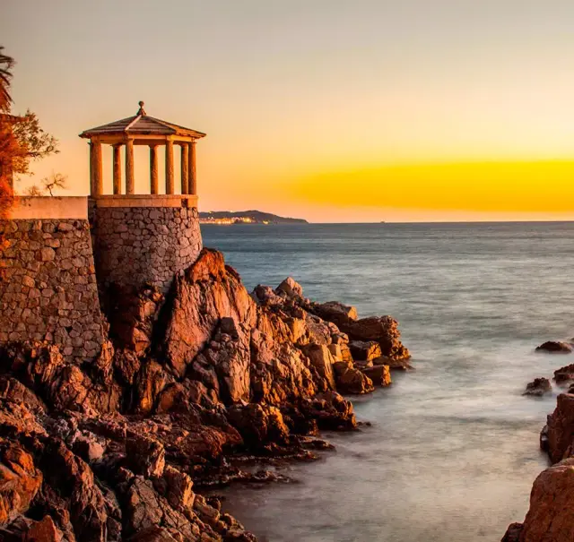 Vista de un mirador de piedra con columnas sobre un acantilado rocoso junto al mar al atardecer, con el cielo en tonos
