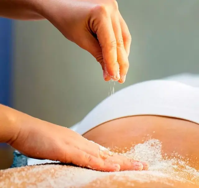 Manos aplicando sal sobre la espalda de una persona tumbada durante un tratamiento de exfoliación corporal en un spa.