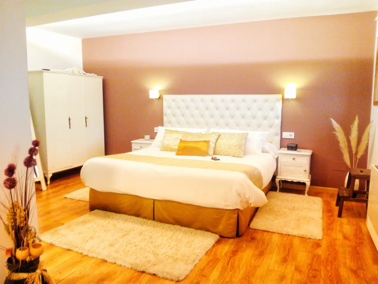 Vista de una suite con cama doble grande con cabecero capitoné blanco, mesitas de noche, armario blanco, suelo