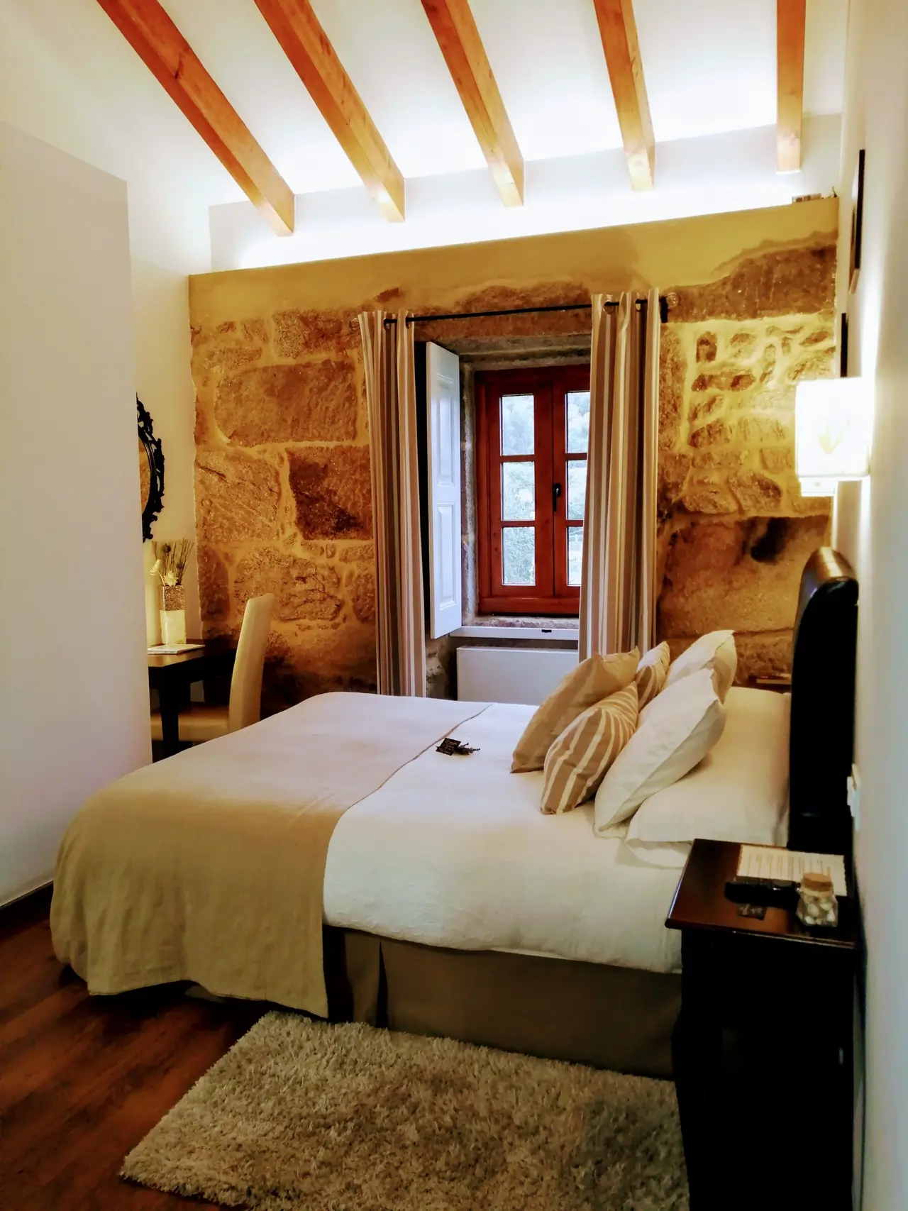 Habitación de suite con cama doble, cabecero oscuro, paredes de piedra vista, techo con vigas de madera, ventana