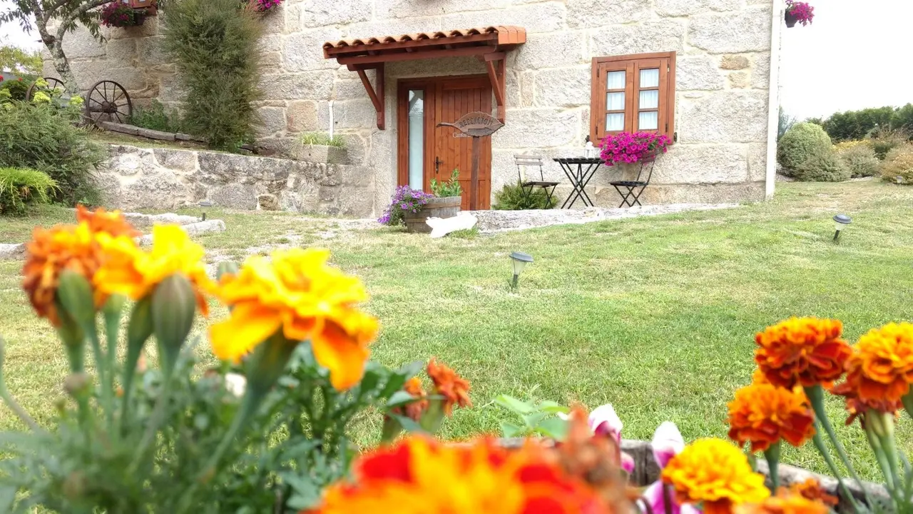 Vista exterior de una casa de piedra con puerta de madera, ventana, mesa y sillas en el jardín, con flores naranjas