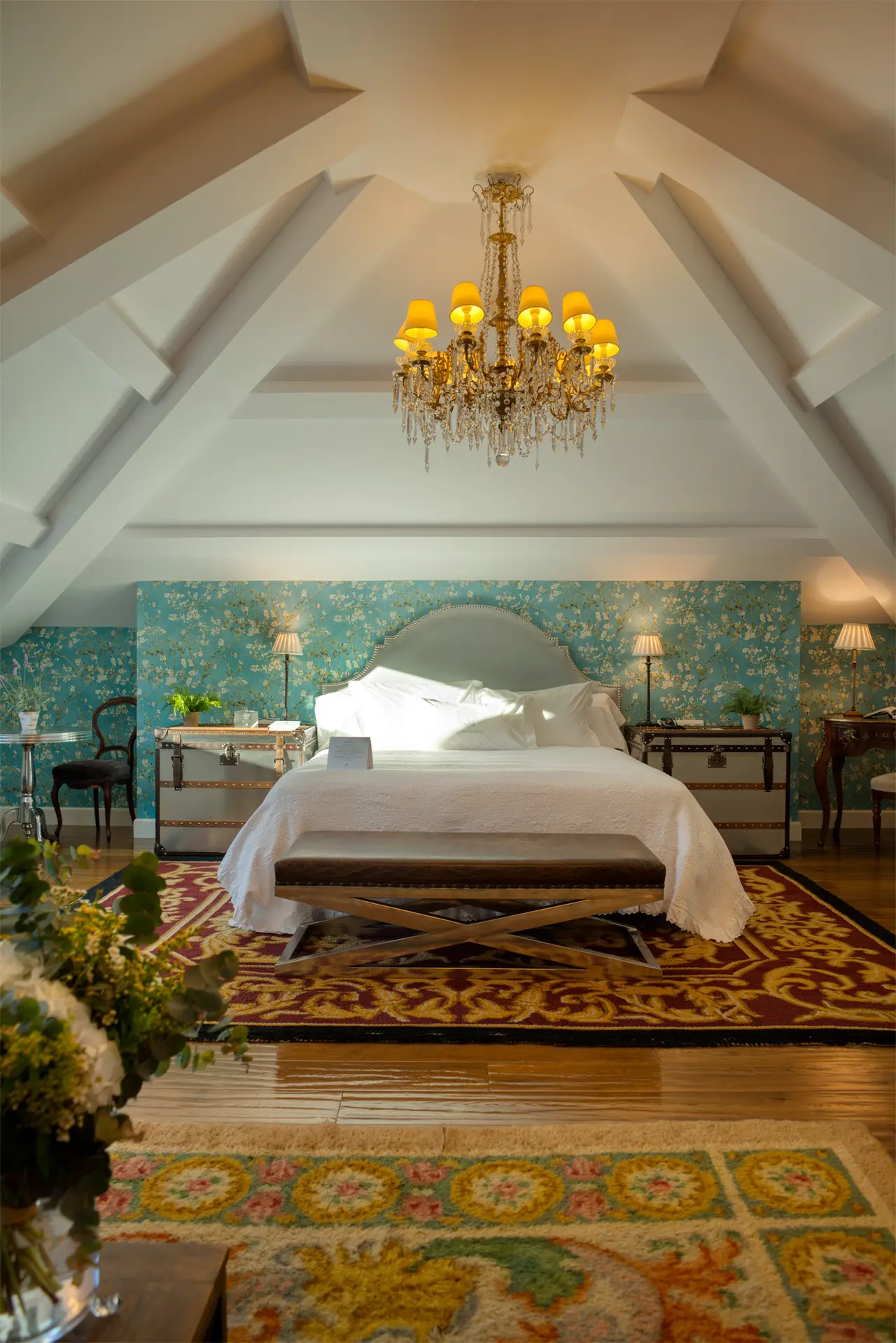 Habitación con techo abuhardillado, cama grande con cabecero gris y ropa de cama blanca, dos mesitas de noche