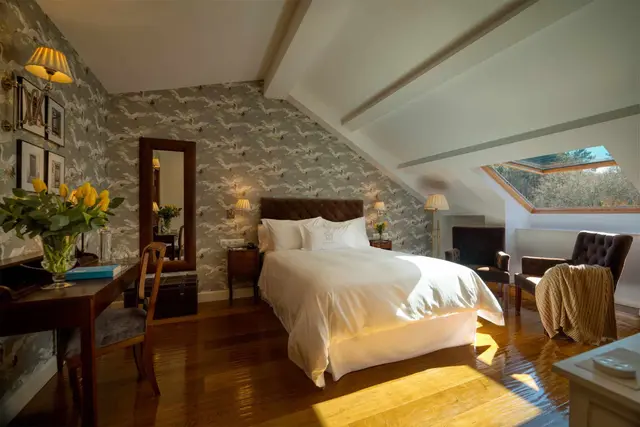 Habitación abuhardillada con cama doble vestida de blanco, escritorio de madera con silla, papel pintado con patrón
