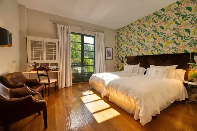 Habitación con cama doble grande vestida de blanco, cabecero oscuro, papel pintado floral en la pared, suelo