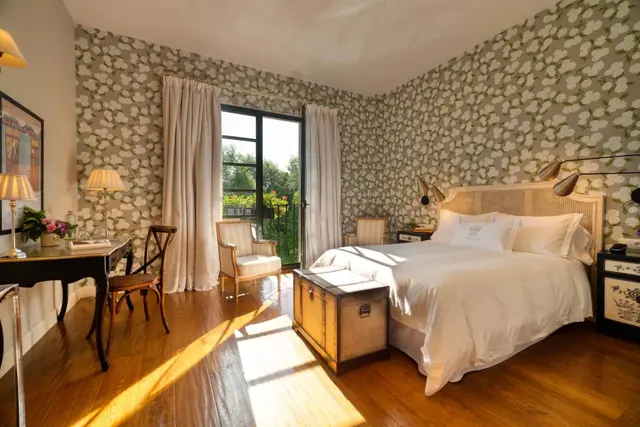 Habitación con cama grande vestida de blanco, papel pintado floral en las paredes, suelo de madera, escritorio