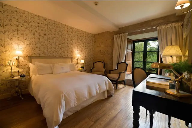 Habitación con cama doble grande vestida de blanco, papel pintado floral en las paredes, suelo de madera, dos sillones