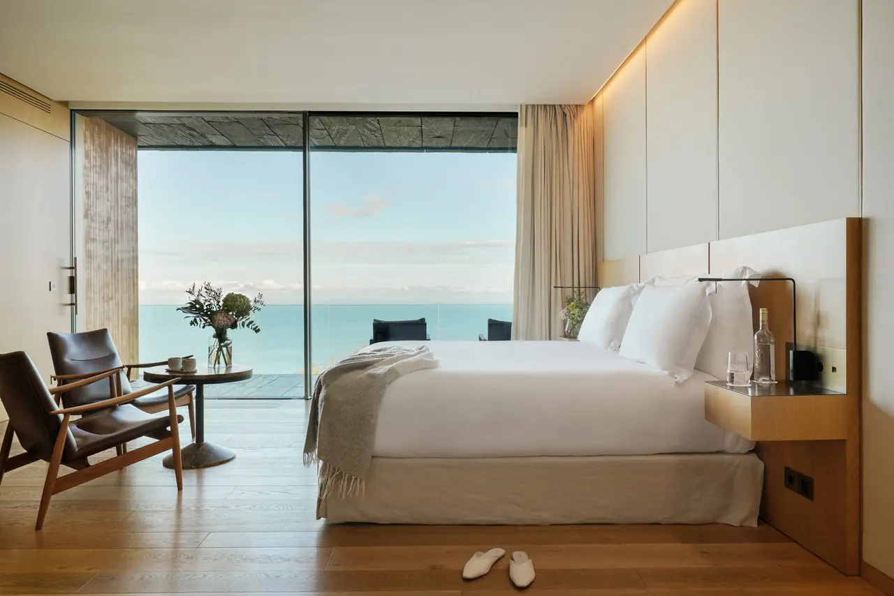 Habitación de hotel con cama grande vestida de blanco, dos sillones de cuero, mesa redonda y gran ventanal abierto