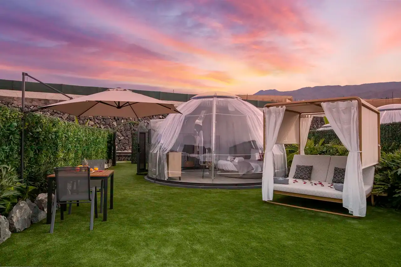 Vista exterior de una suite burbuja transparente con cortinas blancas, situada en un jardín de césped al atardecer.