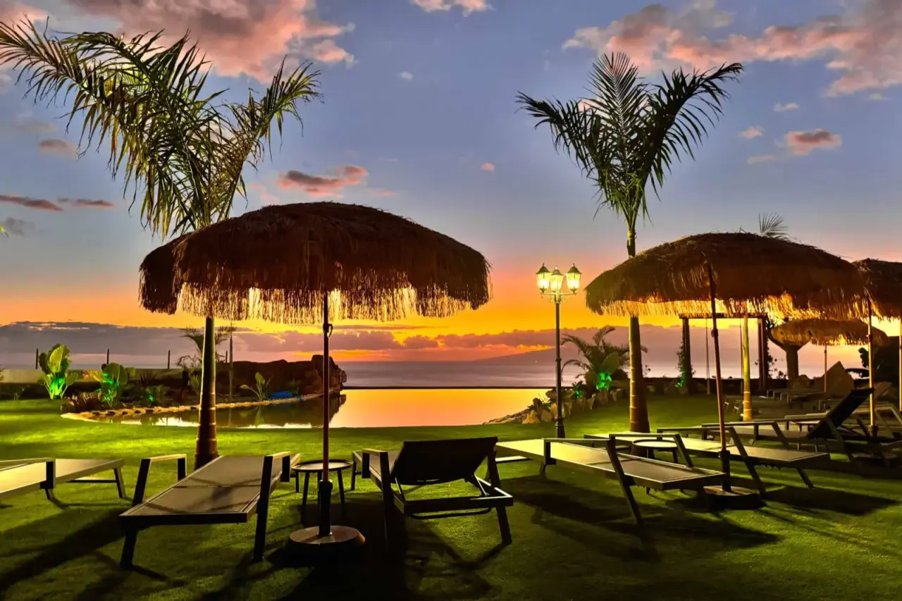 Vista exterior al atardecer de un jardín con césped, palmeras, sombrillas de paja, tumbonas y un farol, con el mar