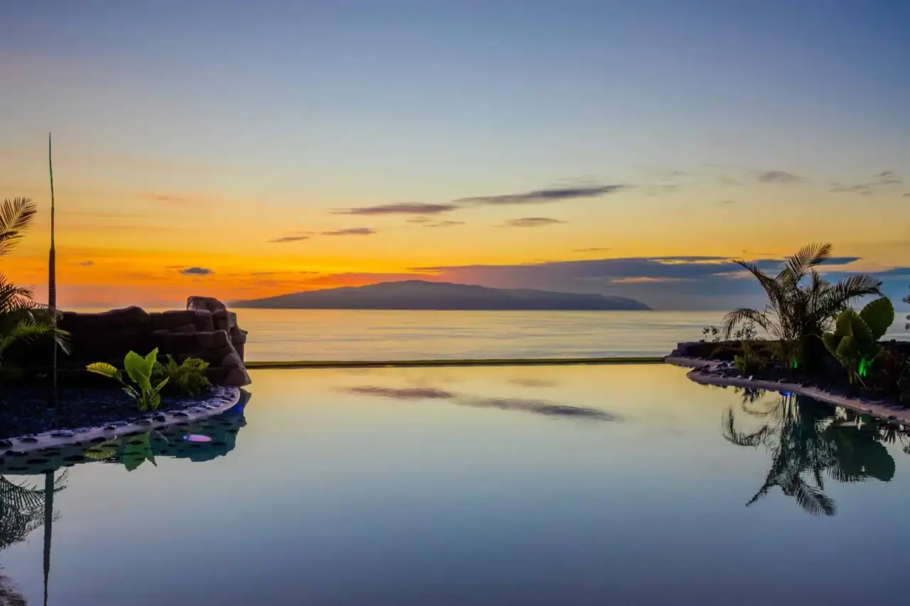 Vista de una piscina infinita con agua calmada reflejando el cielo al atardecer, rodeada de vegetación tropical