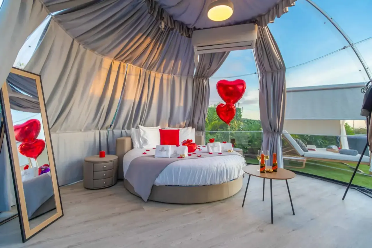 Interior de una suite burbuja con cama redonda central, cortinas de tela gris, globos rojos en forma de corazón, mesa