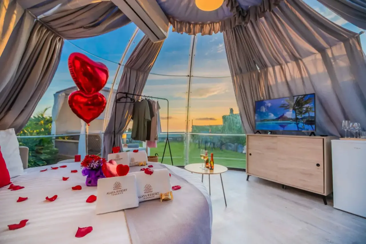 Interior de una suite burbuja con cama redonda decorada con pétalos de rosa y globos en forma de corazón, ventanales