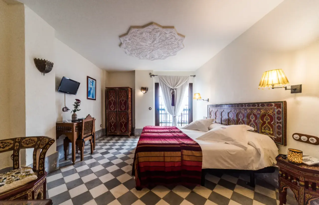 Vista de una habitación de hotel con cama grande vestida con colcha roja y blanca, cabecero decorado, mesitas de noche