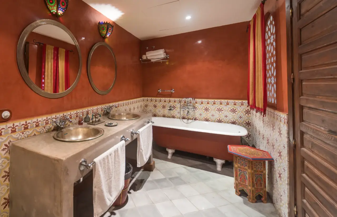 Baño con paredes naranjas y azulejos decorados, doble lavabo de cobre sobre encimera de piedra, toallas blancas