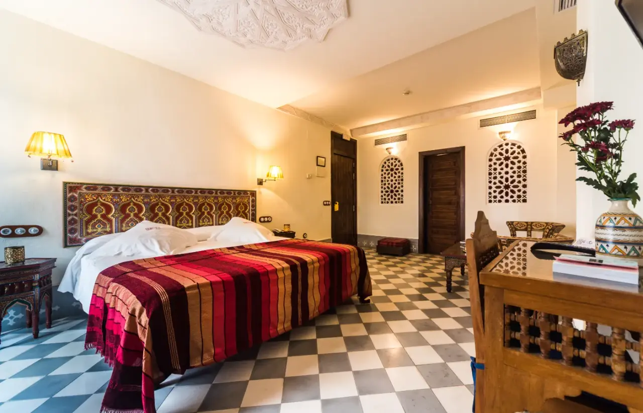 Habitación de hotel con cama grande cubierta por una colcha de rayas rojas, marrones y beige, cabecero decorado