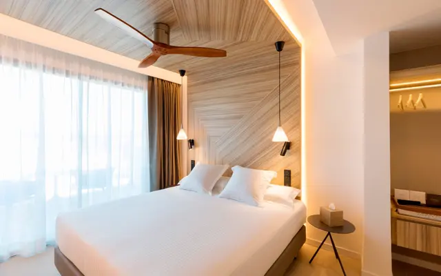 Habitación con cama king size vestida de blanco, cabecero de madera en espiga iluminado, ventilador de techo