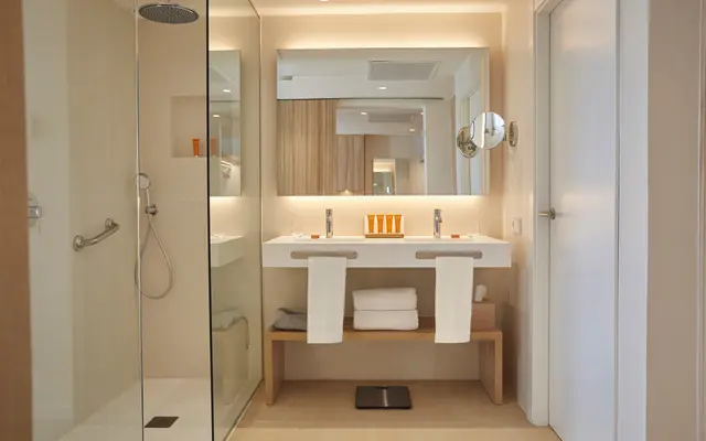 Baño moderno con ducha de lluvia a la izquierda, doble lavabo con grifos, espejo retroiluminado, toallas blancas