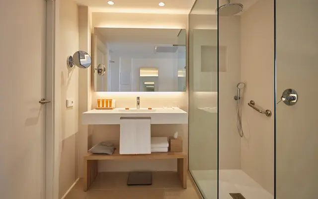 Baño moderno con ducha de cristal, lavabo blanco sobre encimera de madera, espejo retroiluminado, toallas blancas
