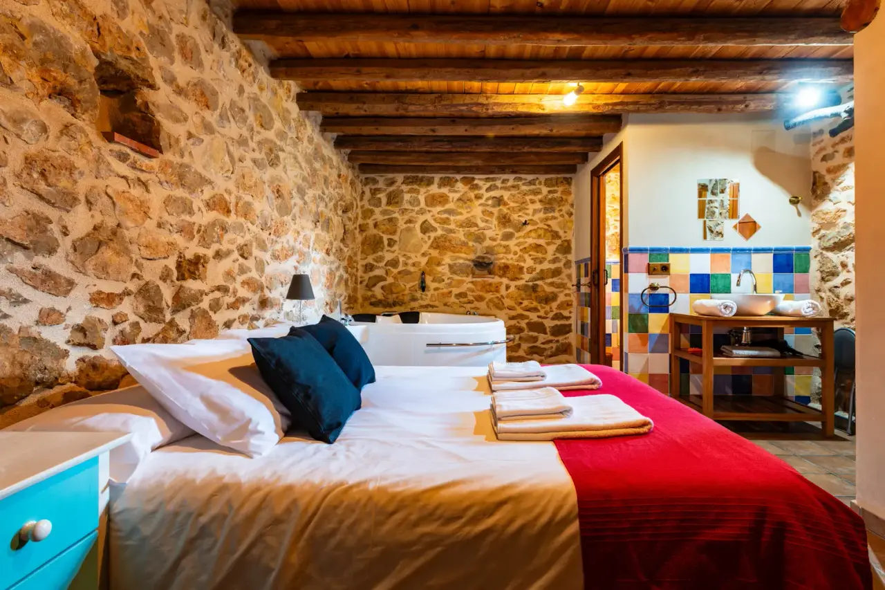 Habitación rústica con cama grande, jacuzzi al fondo y paredes de piedra.