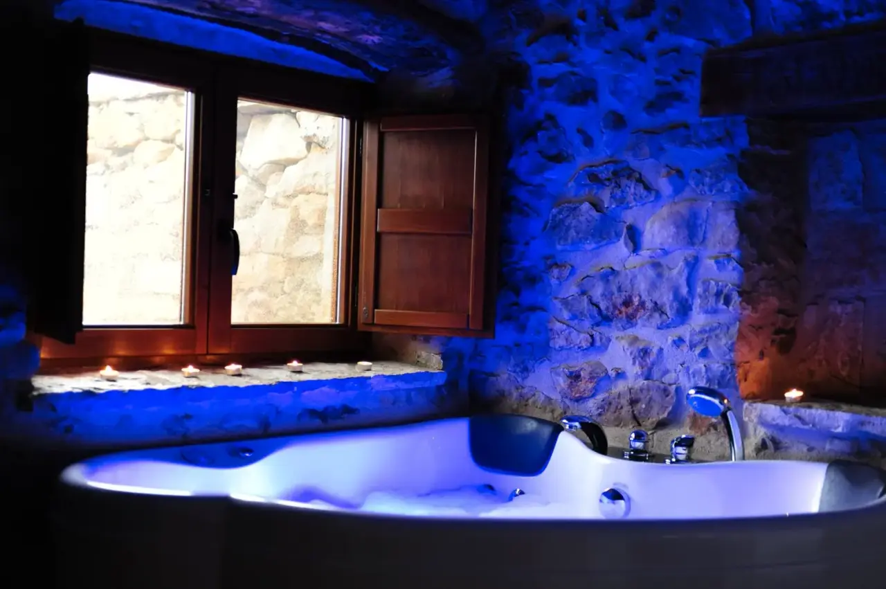 Jacuzzi iluminado en una habitación rústica con paredes de piedra y ventana de madera.