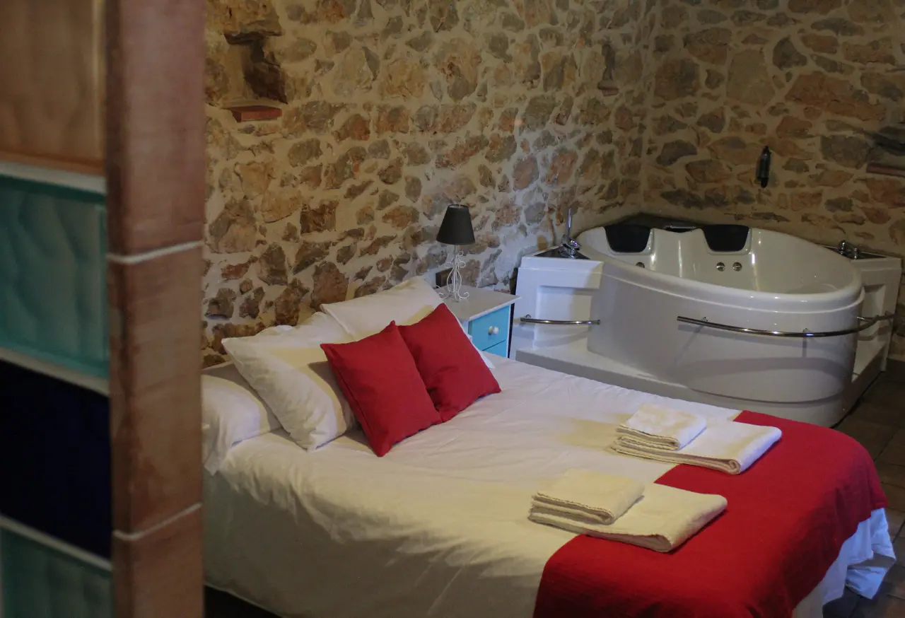 Habitación con cama de matrimonio, paredes de piedra y jacuzzi en la esquina.