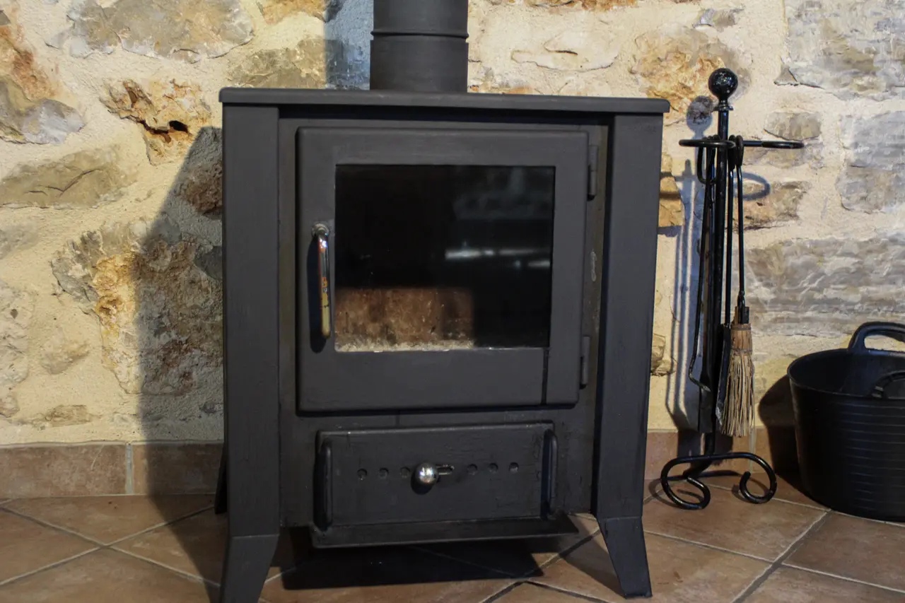 Estufa de leña negra con puerta de cristal y accesorios junto a una pared de piedra rústica.