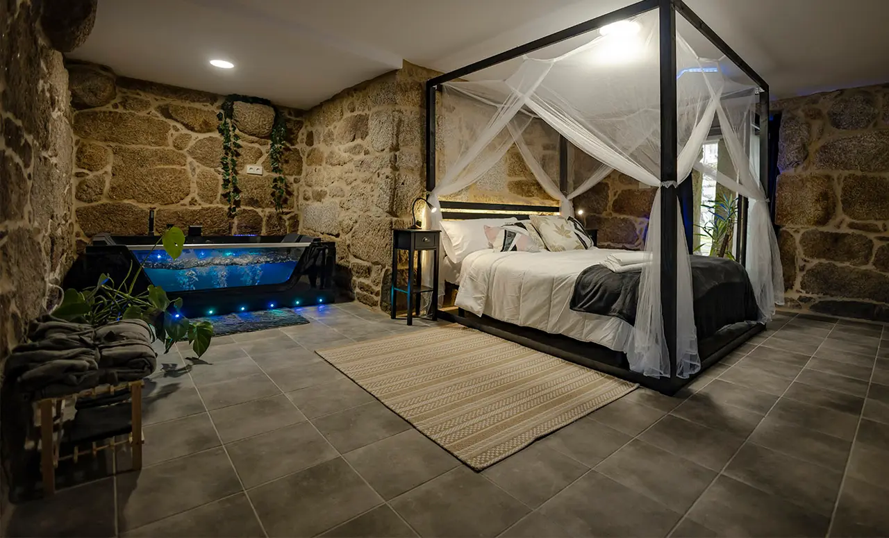 Interior de suite con paredes de piedra, cama con dosel y mosquitero, y jacuzzi iluminado en azul ubicado al fondo