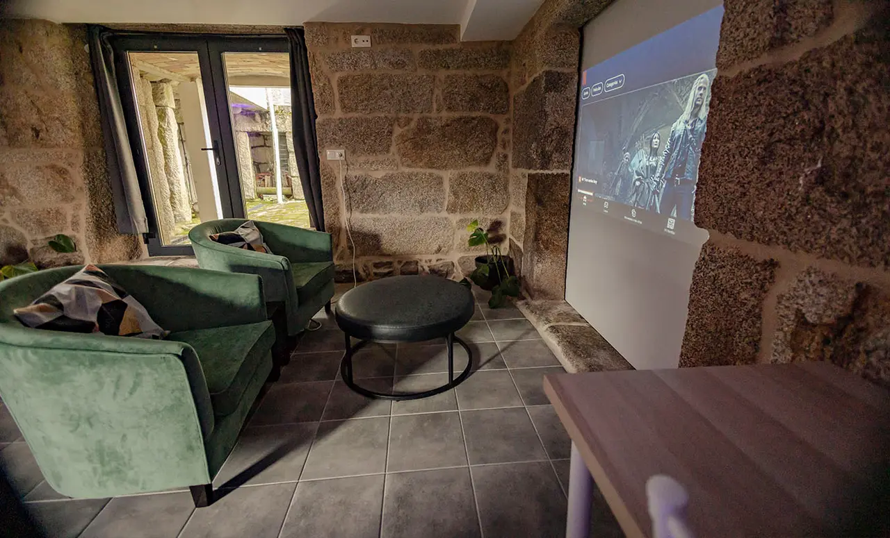 Interior de una habitación con paredes de piedra, dos sillones verdes, una mesa redonda negra, una pantalla