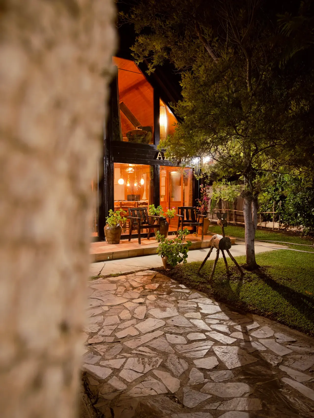 Vista nocturna de la fachada de una cabaña de madera iluminada, con terraza, sillas, macetas y un camino de piedra