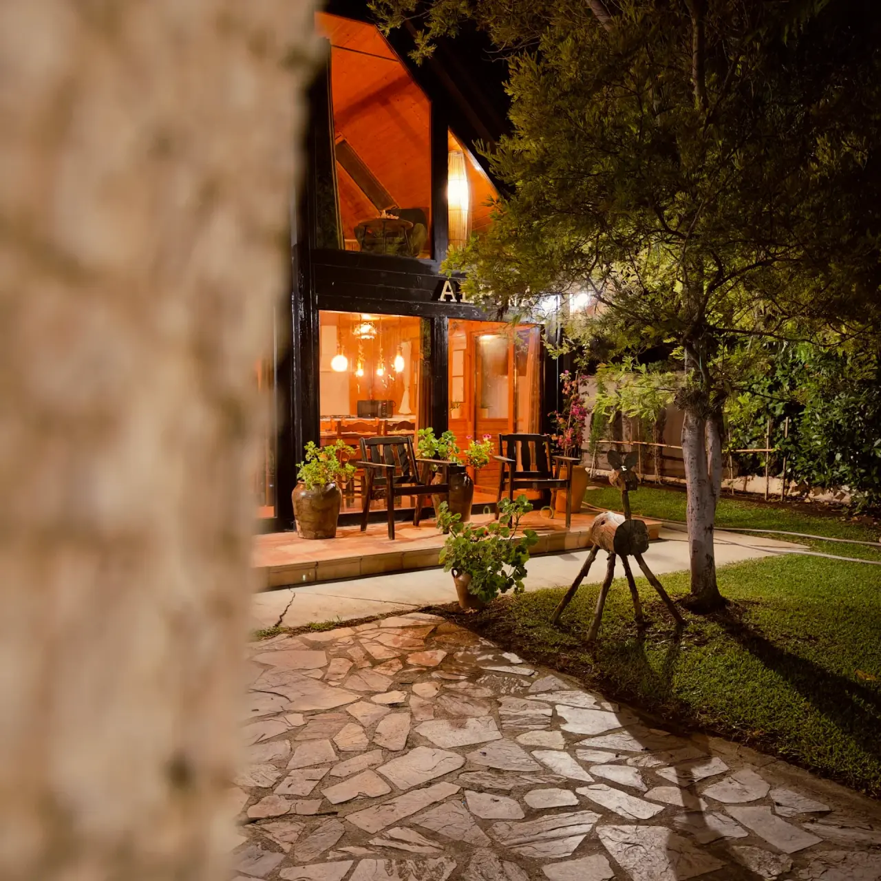 Vista nocturna de la fachada de una cabaña de madera iluminada, con terraza, sillas, macetas y un camino de piedra