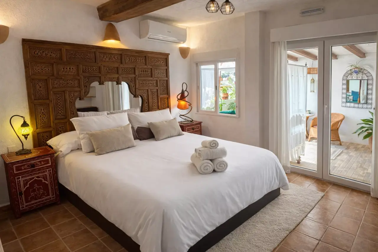 Habitación con cama grande, cabecero de madera tallada, terraza con puertas de cristal y suelo de terracota.