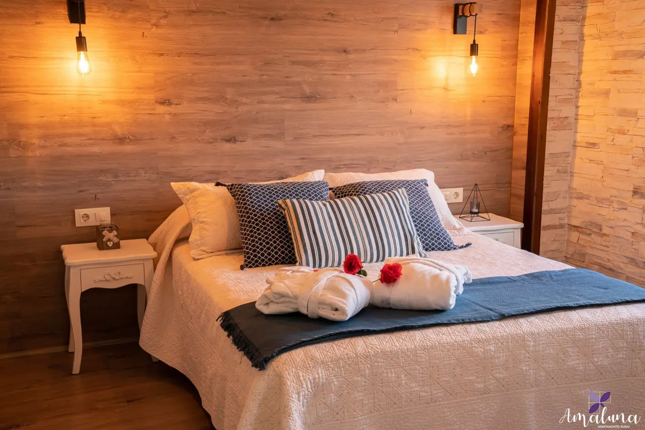 Cama matrimonial con cojines decorativos, toallas enrolladas con flores y manta azul en una habitación con paredes