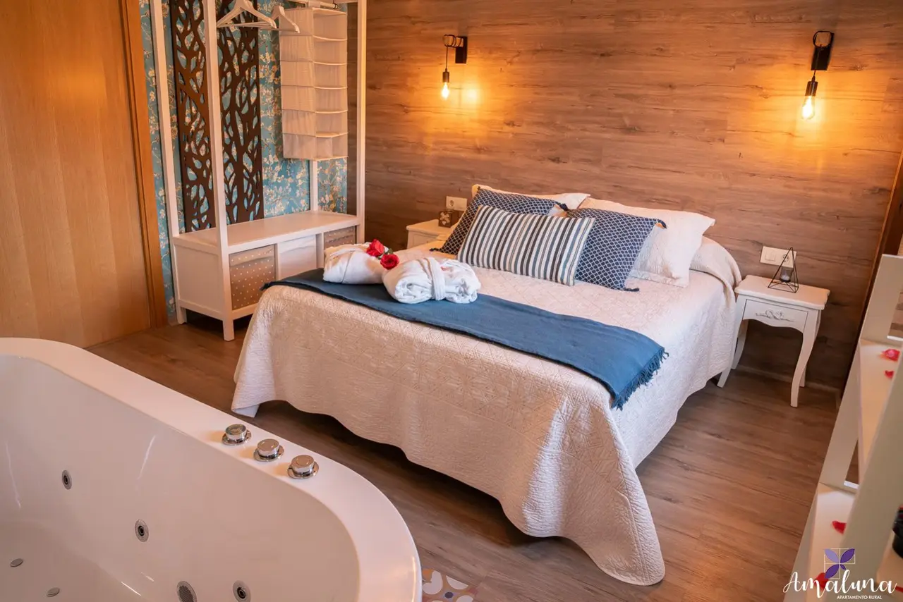 Habitación con cama grande, bañera de hidromasaje en primer plano y paredes de madera clara.