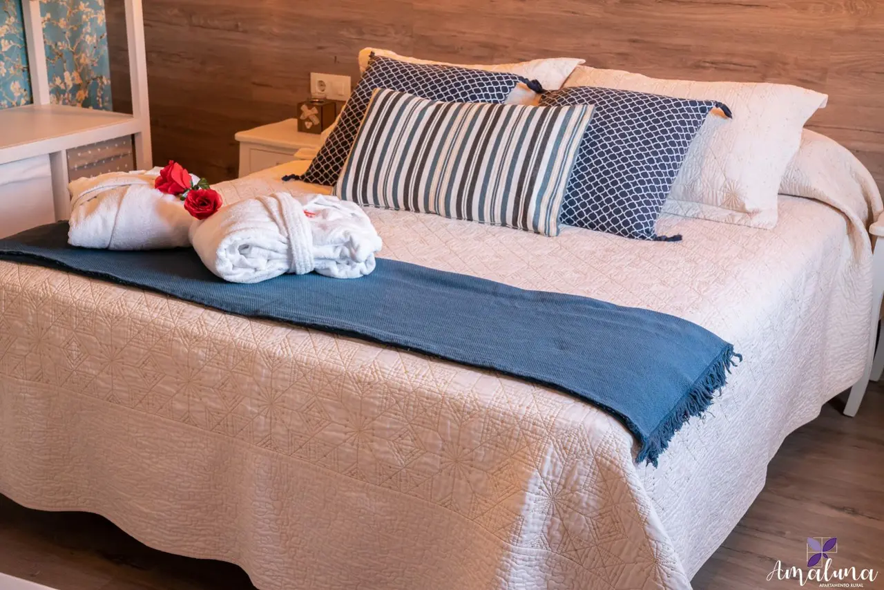 Cama matrimonial en habitación con colcha clara, cojines decorativos y toallas enrolladas con rosas rojas
