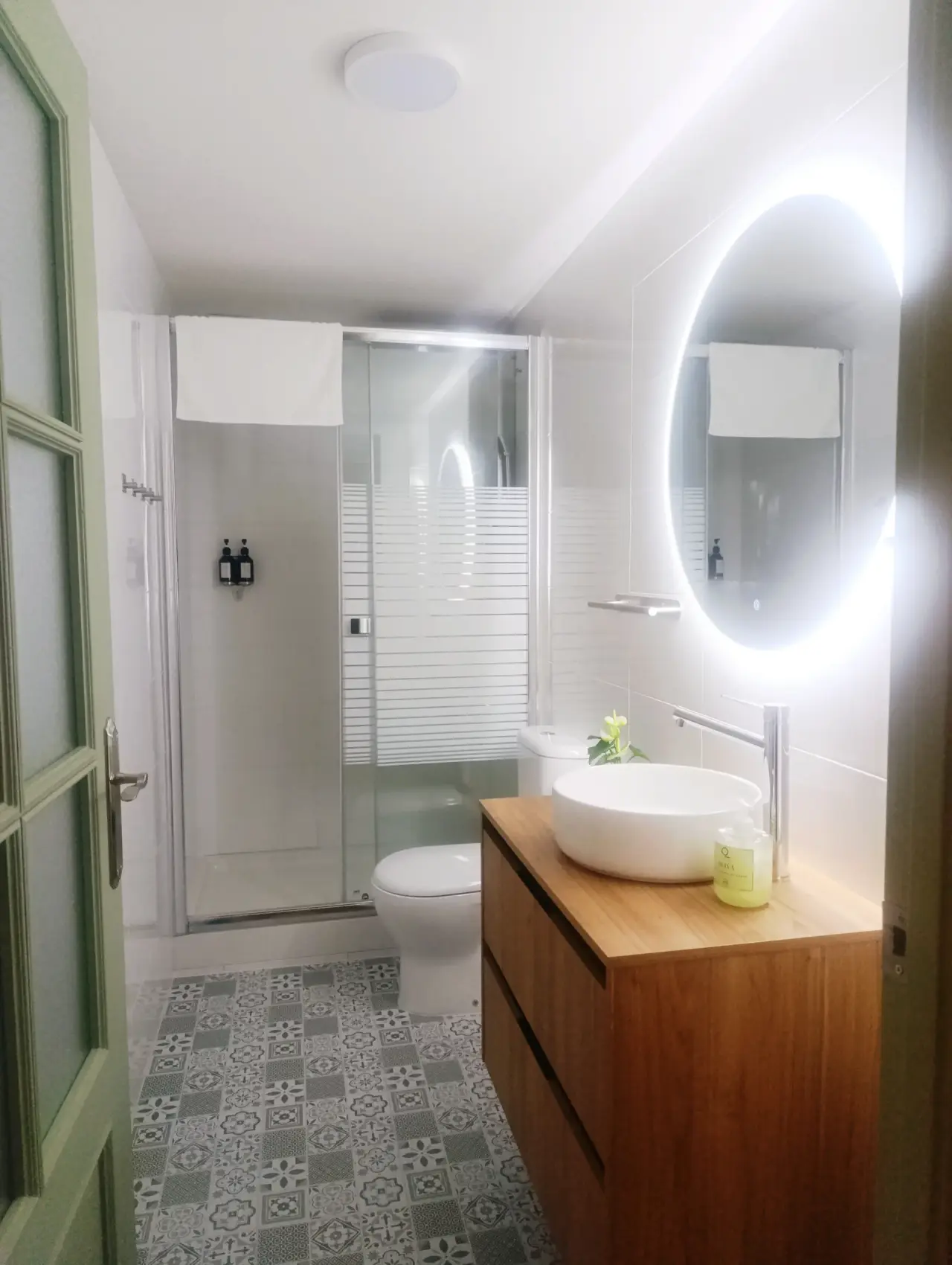 Baño moderno con ducha de cristal, inodoro, lavabo redondo sobre mueble de madera, gran espejo circular iluminado