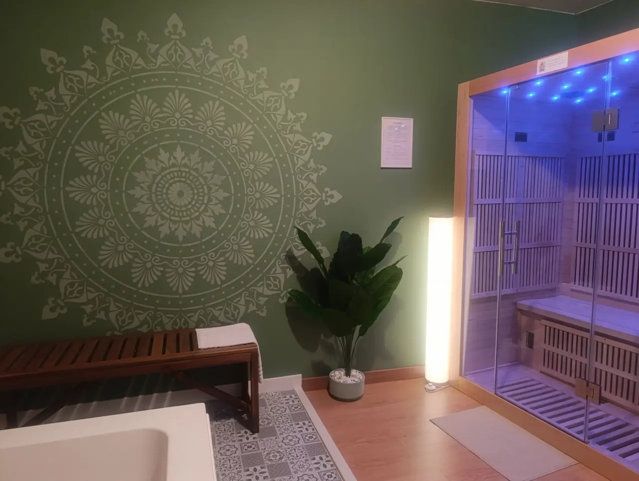 Interior de un spa con una pared verde decorada con un gran mandala blanco, un banco de madera con toalla, una planta