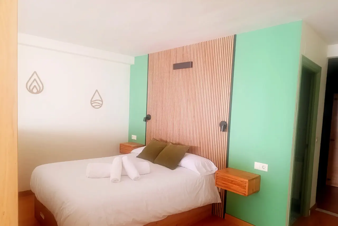 Habitación con cama doble vestida de blanco, dos almohadas verdes, toallas enrolladas, cabecero de madera vertical
