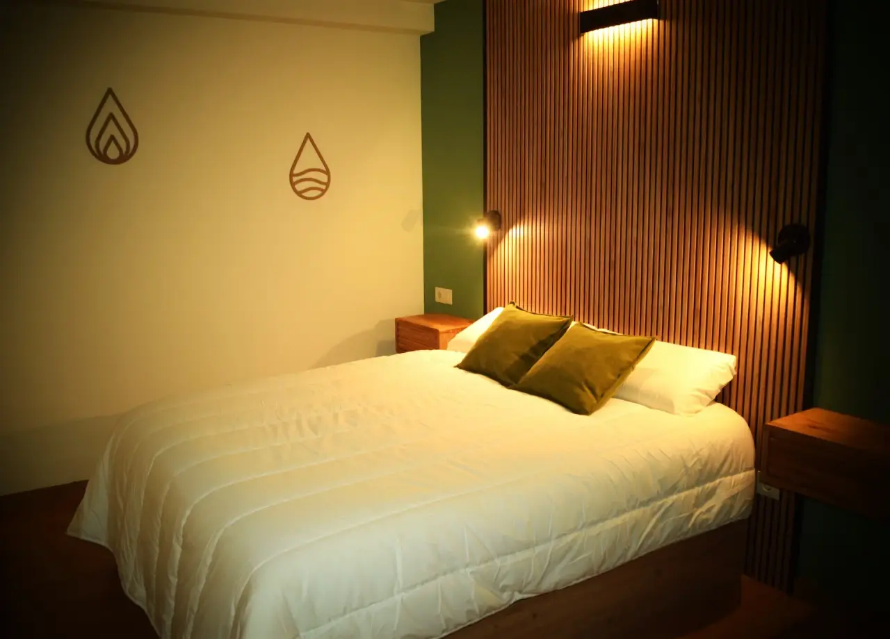 Habitación con cama doble grande vestida de blanco, cabecero de madera vertical iluminado por apliques de pared