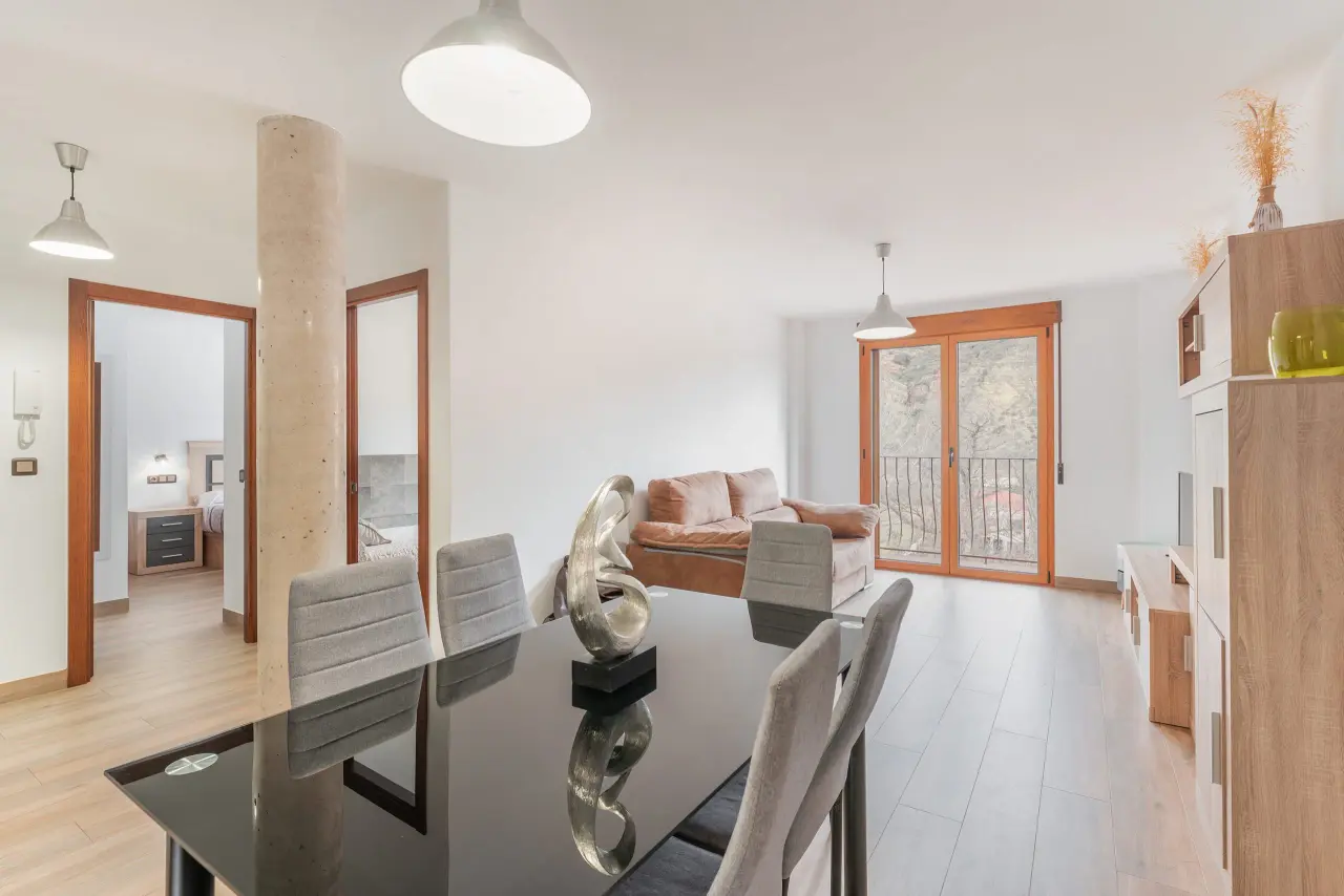 Interior de un apartamento con comedor en primer plano, mesa negra con sillas grises, sofá en el fondo y puertas