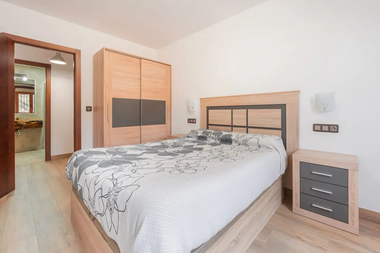 Habitación con cama doble, armario empotrado de madera, mesita de noche y puerta abierta que da a un baño con lavabo