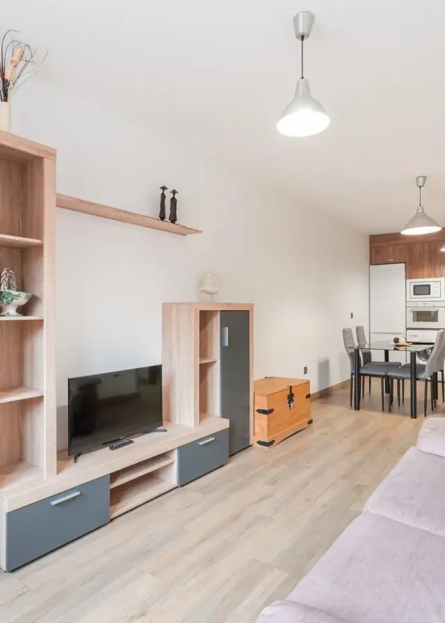 Interior de un salón con mueble de televisión en madera clara y gris, sofá en primer plano, mesa de comedor con sillas