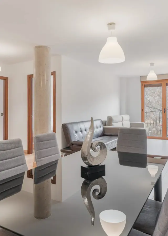Interior de un salón comedor con mesa de cristal, sillas tapizadas en gris, sofá de cuero, columna central