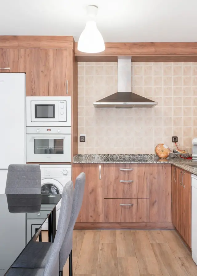 Vista de una cocina moderna con muebles de madera, encimera de granito, campana extractora, horno, microondas