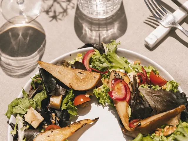Plato de ensalada gourmet con pera asada, tomates cherry, queso y nueces sobre mesa con cubiertos y vasos.