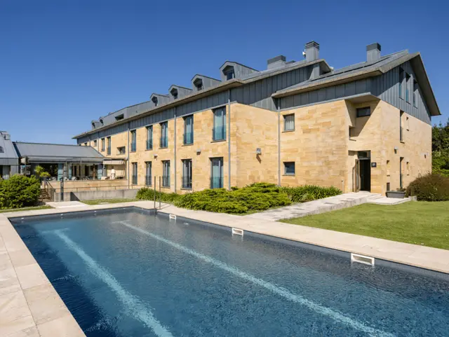 Piscina rectangular al aire libre frente a un edificio de piedra con múltiples ventanas bajo un cielo azul.