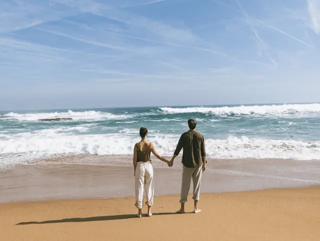 Pareja de espaldas tomada de la mano mirando el mar desde la playa.