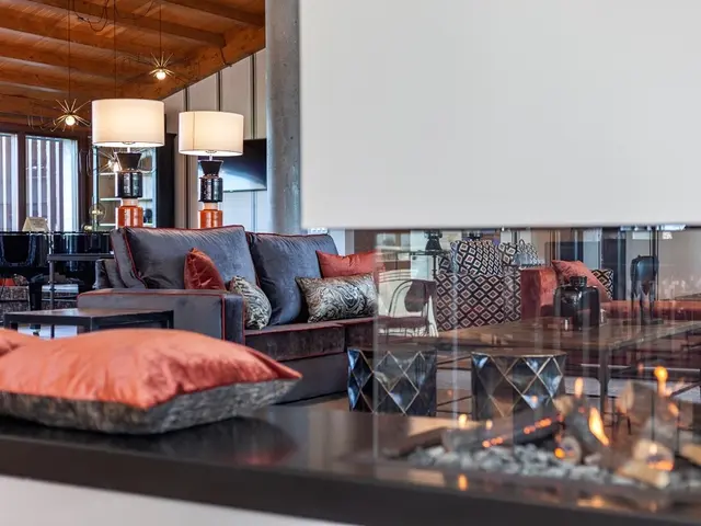 Salón con sofá azul, cojines naranjas y chimenea encendida en primer plano, bajo techo de madera y vigas vistas.