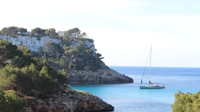 Vista panorámica de una cala con agua turquesa, un velero anclado y un acantilado blanco con vegetación y edificios