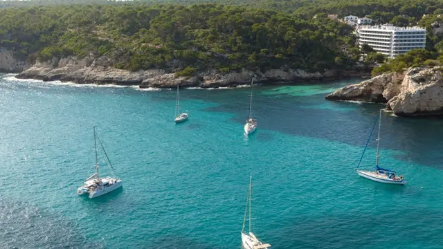 Vista aérea de una cala con agua turquesa y varios veleros anclados, rodeada de acantilados rocosos y vegetación