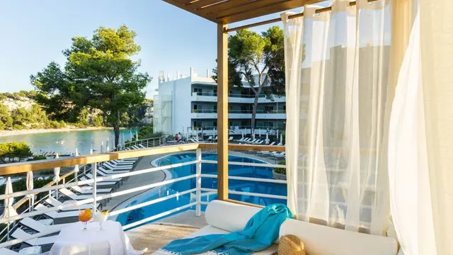 Vista desde una terraza con cortinas blancas hacia una piscina curva y un jardín con árboles, con una mesa con bebida
